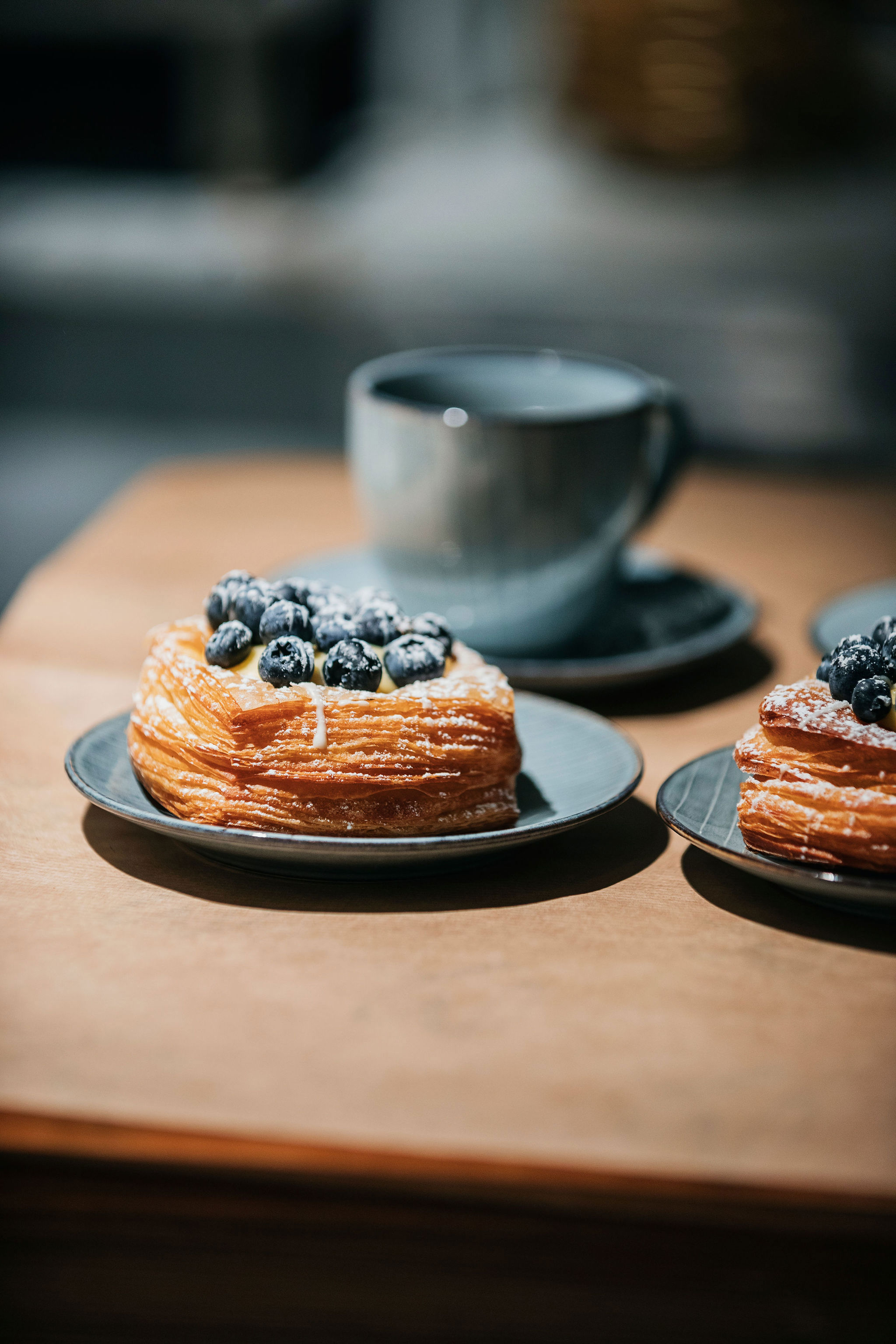Närbild på en blå kaffekopp och ett fat med en bakelse toppad med färska blåbär. 