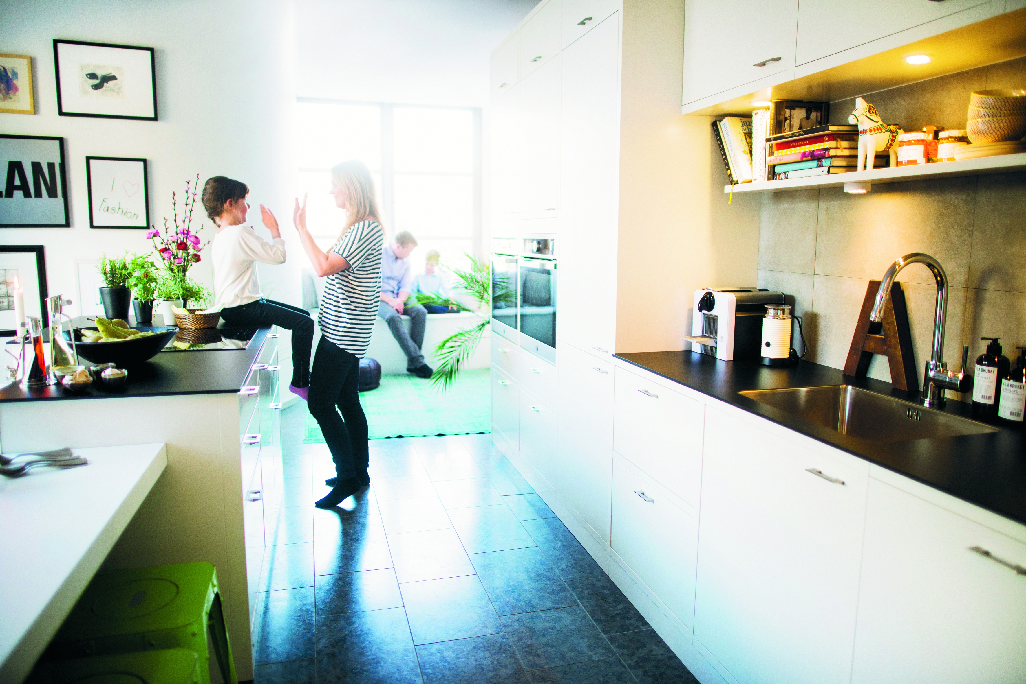 En mamma leker med sitt barn som sitter på en köksbänk i ett vitt öppet kök med en pappa och ett barn lite suddigt i bakgrunden. 