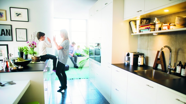 En mamma leker med sitt barn som sitter på en köksbänk i ett vitt öppet kök med en pappa och ett barn lite suddigt i bakgrunden. 