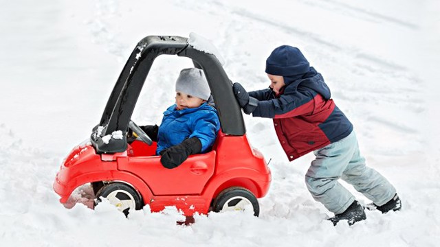 ett litet barn skjuter på ett annat barn som sitter i en liten leksaksbil omringade av mycket snö. 