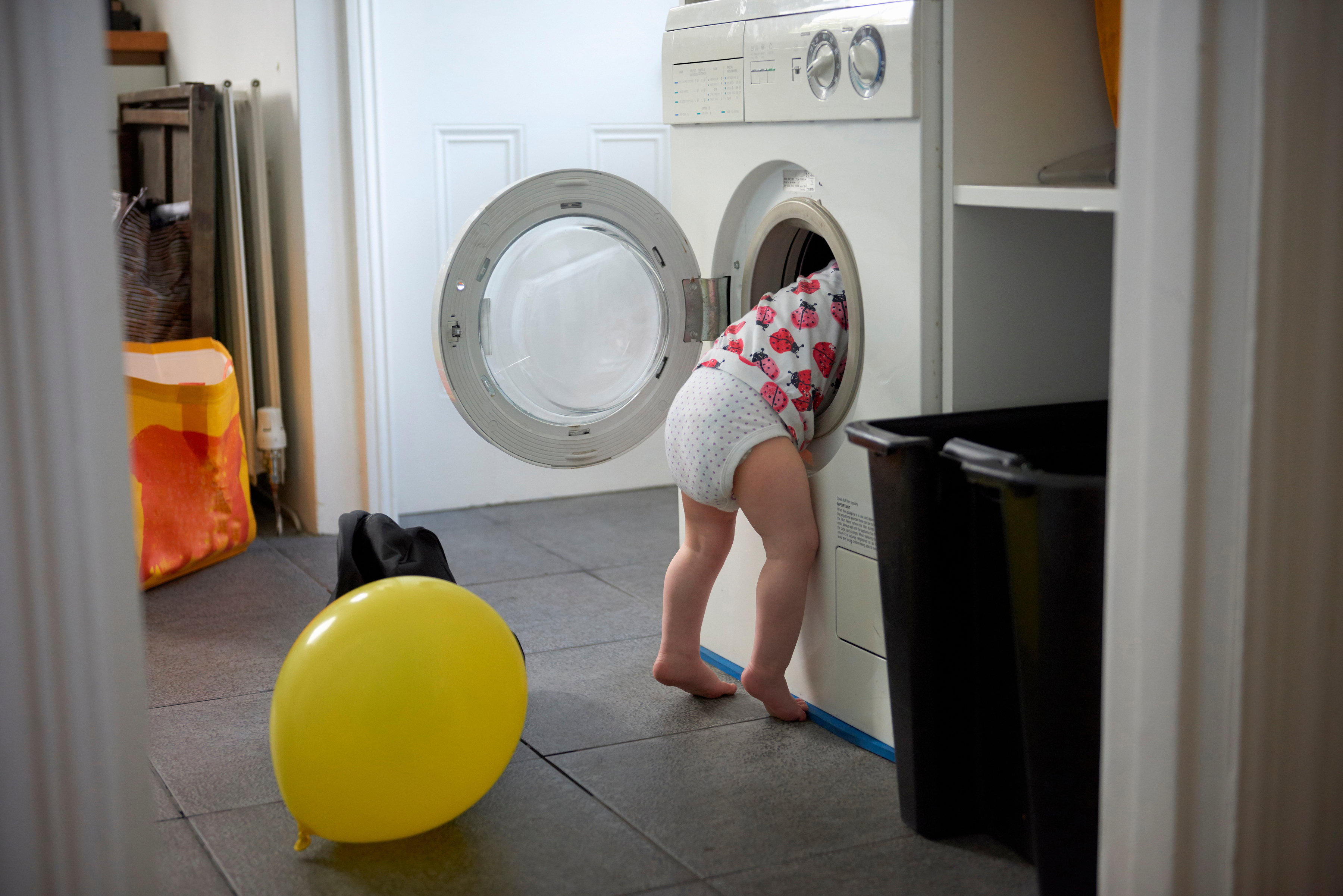 Ett badrum med en tvättmaskin i fokus där ett litet blöjbarn kikar in med hela överkroppen i tvättmaskinenslucka. Några ballonger ligger på golvet bredvid. 