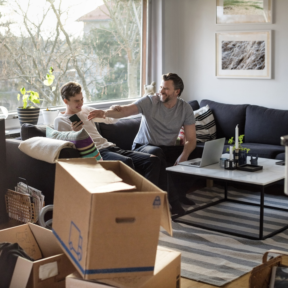 En pappa och hans son busar i soffan som står i ett vardagsrum med flyttkartonger. 