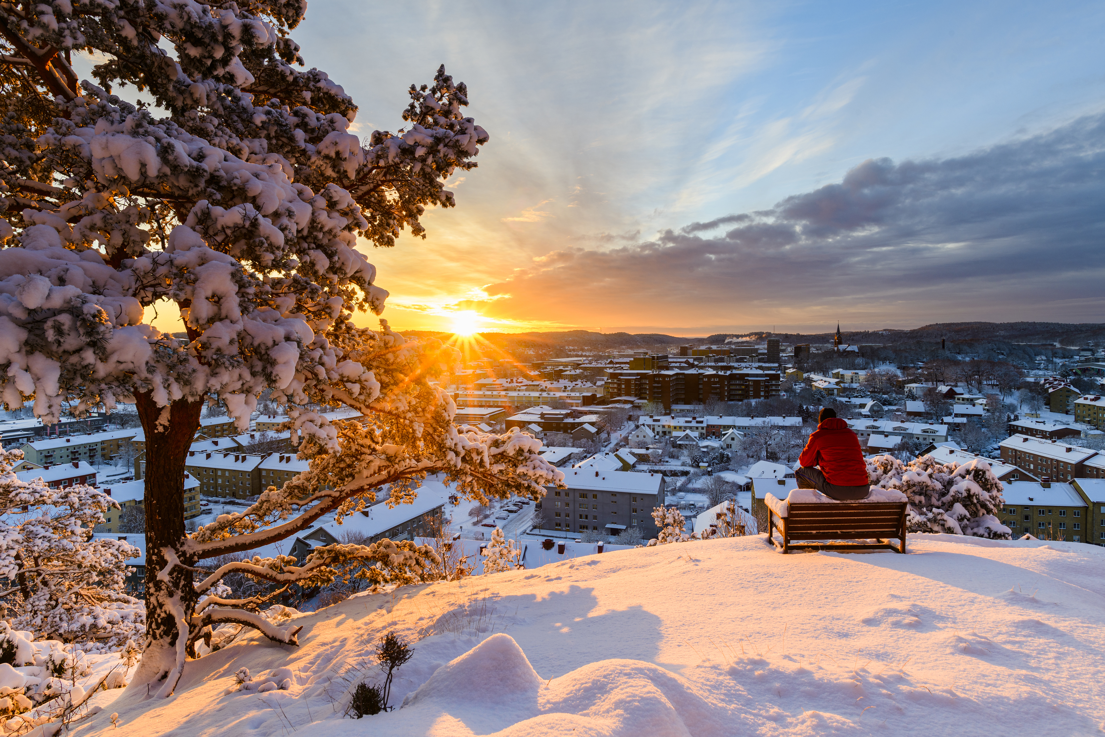 En person sitter på ett högt berg omgiven av snö och blickar ut över Mölndal i solnedgången.