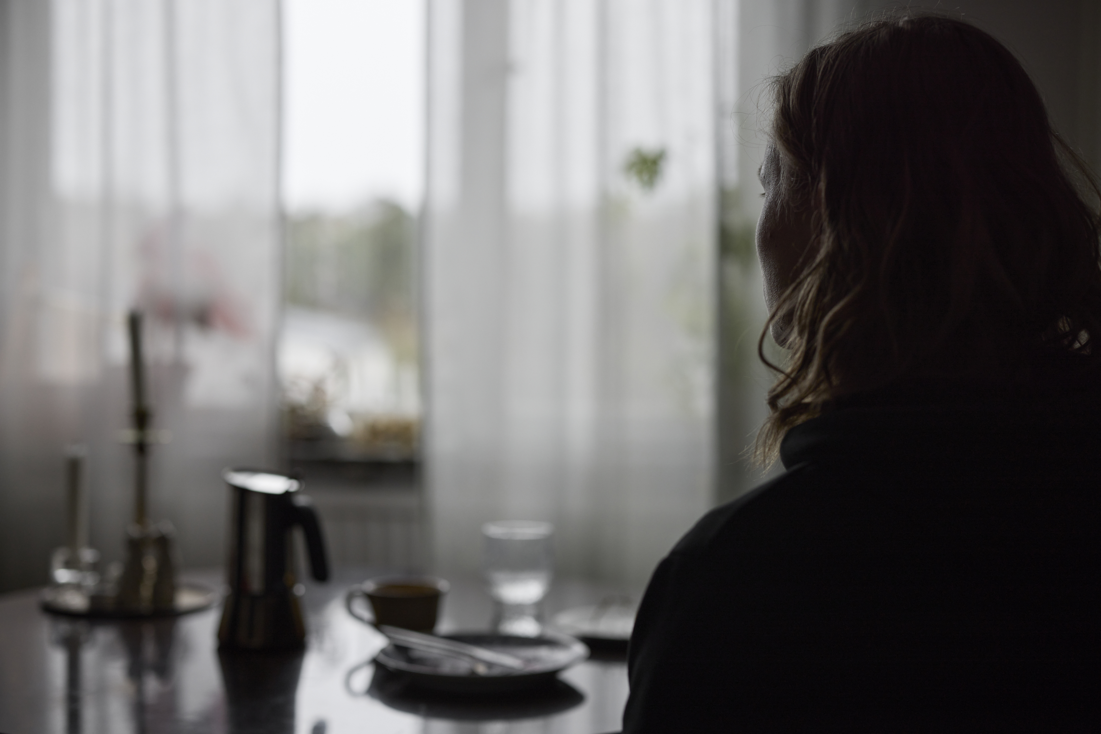 Närbild på en person med axel-långt hår i en mörk siluett som sitter ensam vid ett matbord och vemodigt tittar ut genom fönstret. 